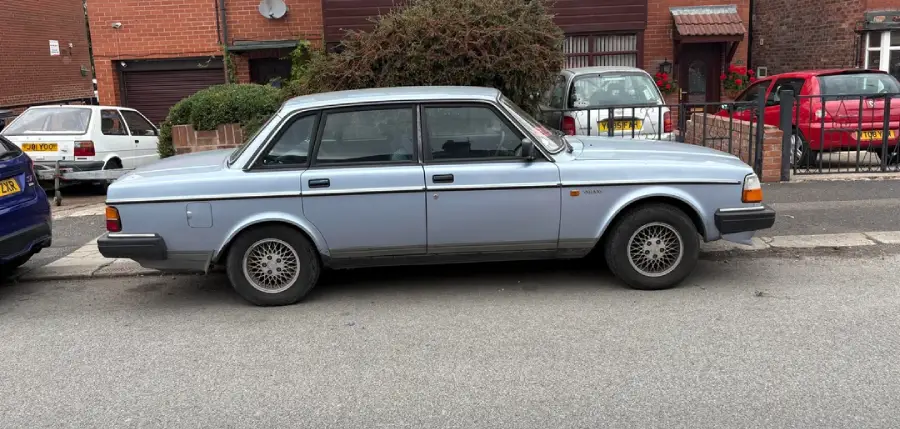A light blue classic sedan is parked on the street in front of a brick house and bushes, with other cars and buildings visible in the background.