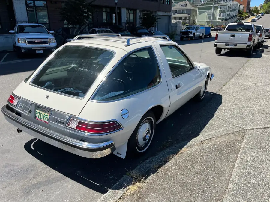 A white vintage AMC Pacer car is parked on a street near a curb, with other vehicles and houses visible in the background on a sunny day.