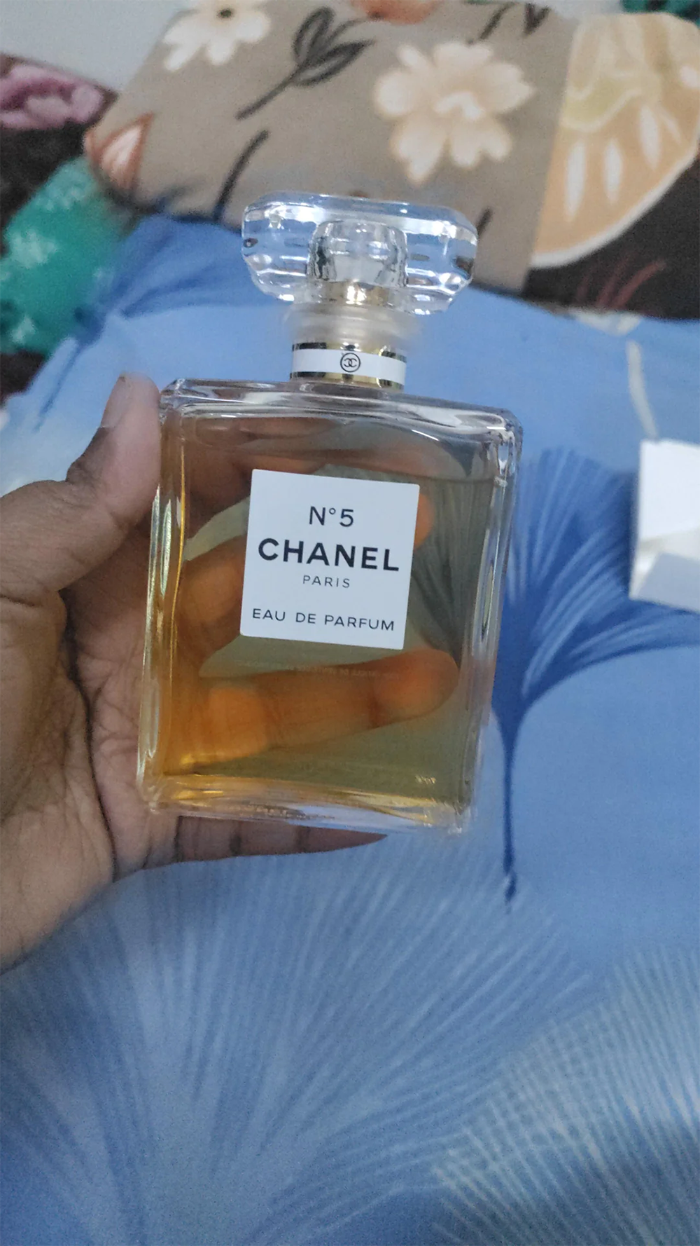 A hand holds a bottle of Chanel No. 5 Eau de Parfum against a blue surface with a floral-patterned pillow in the background.