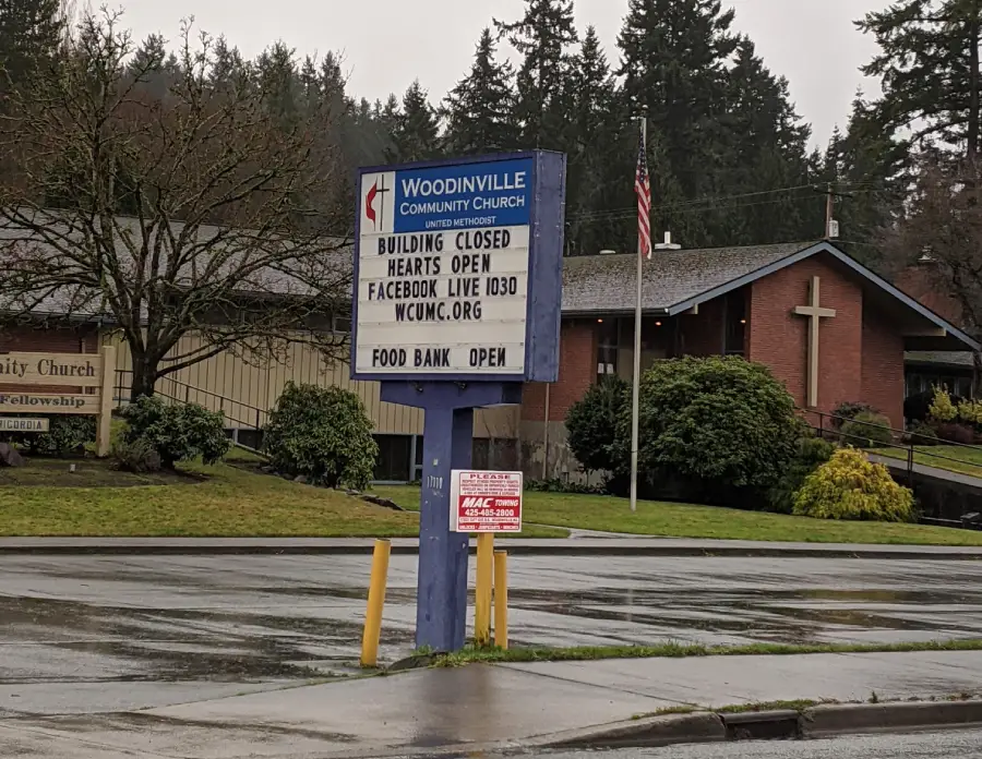 A sign outside Woodinville Community Church reads: “Building closed, hearts open. Facebook live 10:30. wcmc.org. Food bank open.” Church building and trees are in the background on a rainy day.