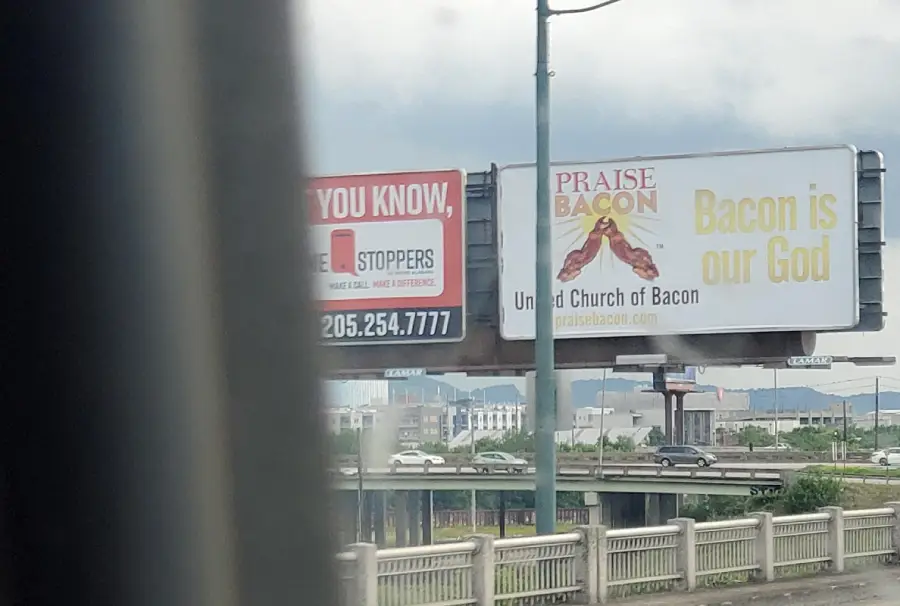 A billboard reads “Praise Bacon. Bacon is our God. United Church of Bacon” with bacon strips shown. Another partially visible sign is next to it, viewed from a moving vehicle above a city street.