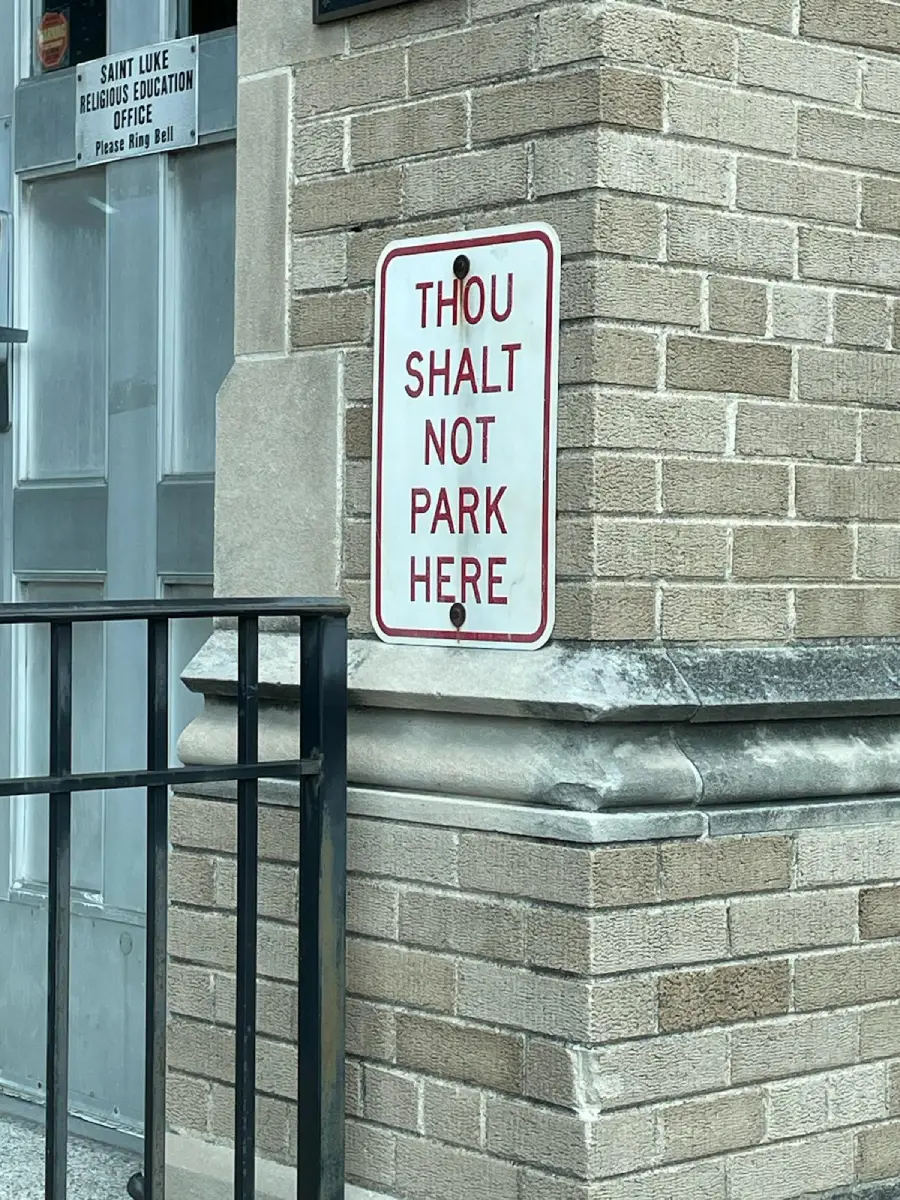 A white sign on a brick wall reads “THOU SHALT NOT PARK HERE” in red letters, next to a door labeled “Saint Luke Religious Education Office.” A black metal railing is in the foreground.