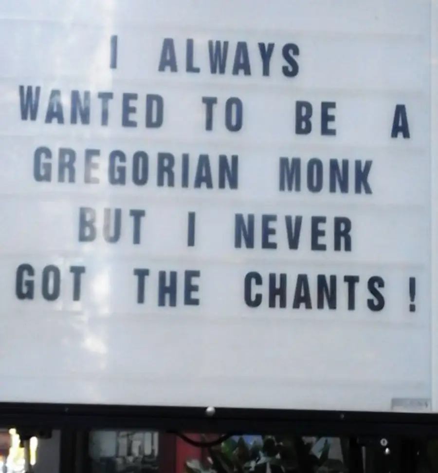 A sign reads: “I always wanted to be a Gregorian monk but I never got the chants!” The message uses wordplay, replacing “chance” with “chants.”
