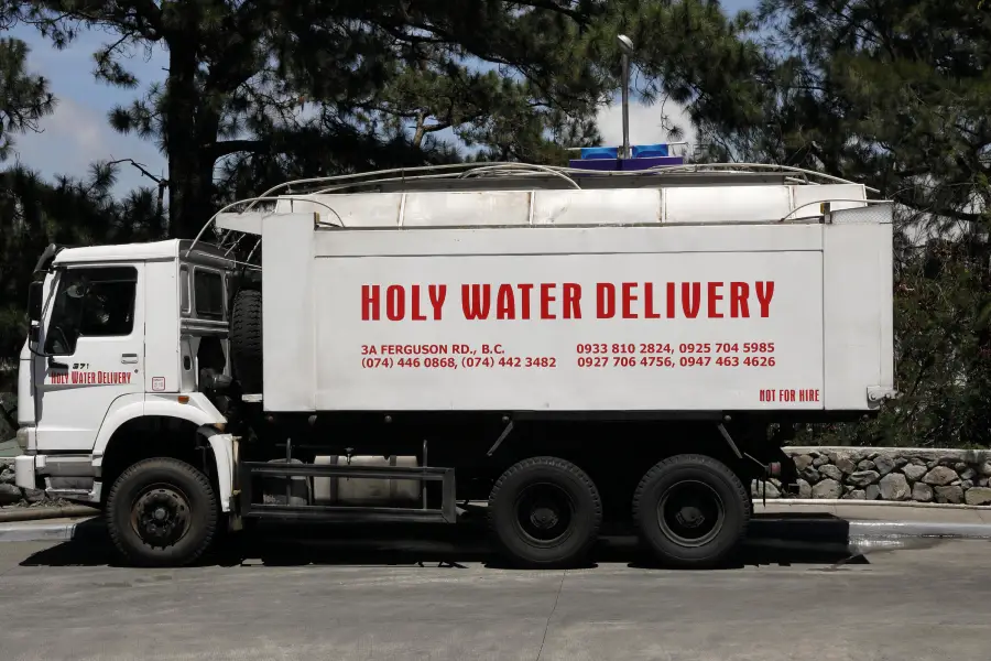 A white tanker truck labeled “Holy Water Delivery” with contact numbers and address, parked on a sunny road near trees and a stone wall.