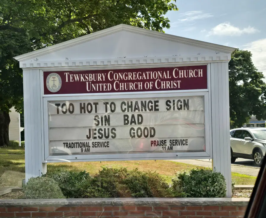 A church sign reads: “TOO HOT TO CHANGE SIGN. SIN BAD. JESUS GOOD.” Service times and the church name are also displayed. Green trees and a car are in the background.