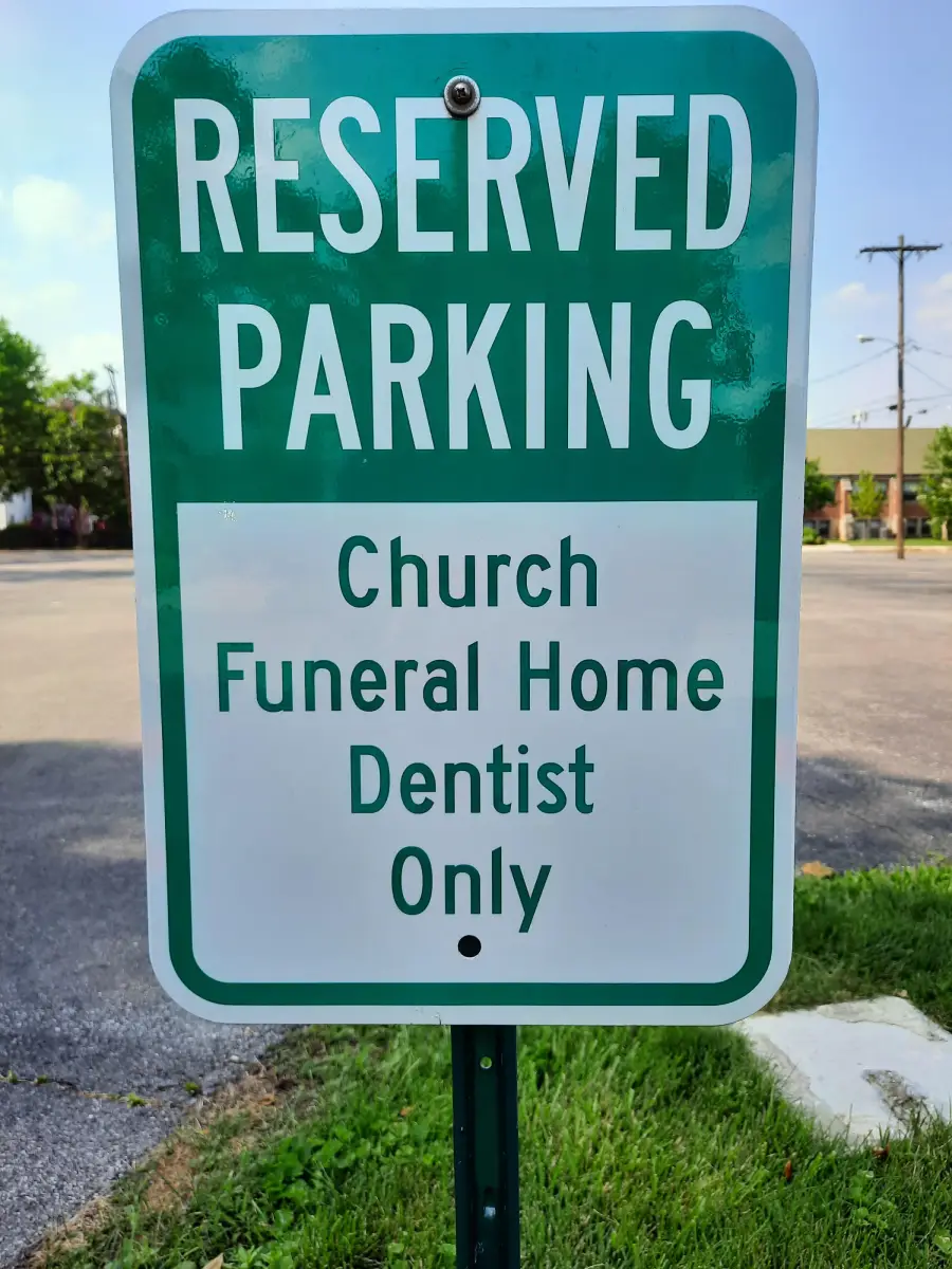 Green and white sign reads “Reserved Parking. Church Funeral Home Dentist Only,” standing on a grassy area near a parking lot with trees and buildings in the background.