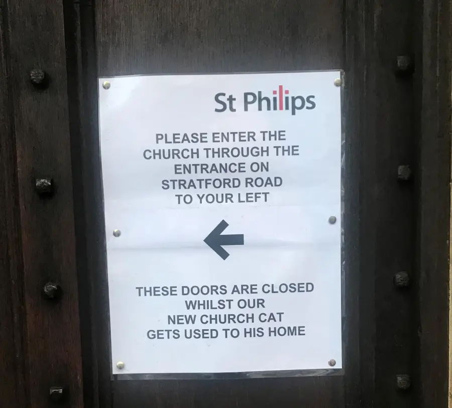 A sign on a wooden door reads: “St Philips. Please enter the church through the entrance on Stratford Road to your left. These doors are closed whilst our new church cat gets used to his home,” with an arrow pointing left.
