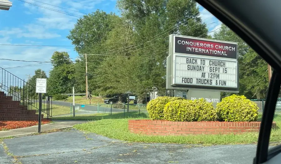 A church sign reads: “Back to Church Sunday Sept 15 at 12PM Food Trucks & Fun.” The building and sign are surrounded by bushes, trees, and a parking lot, viewed from a nearby car.