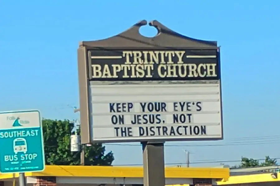 A church sign reads: “KEEP YOUR EYE’S ON JESUS. NOT THE DISTRACTION.” The sign is for Trinity Baptist Church, and a bus stop sign is visible beside it.