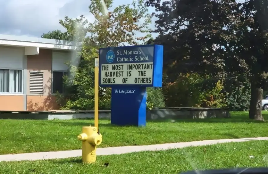 A sign outside St. Monica’s Catholic School reads, “THE MOST IMPORTANT HARVEST IS THE SOULS OF OTHERS.” A yellow fire hydrant is on the grass in front of the sign. Trees and part of a building are visible nearby.