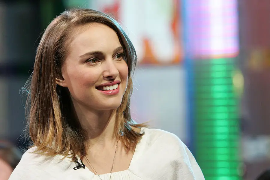 A woman with shoulder-length brown hair and a white top smiles, looking slightly to the side. The background is blurred with colorful lights and shapes.