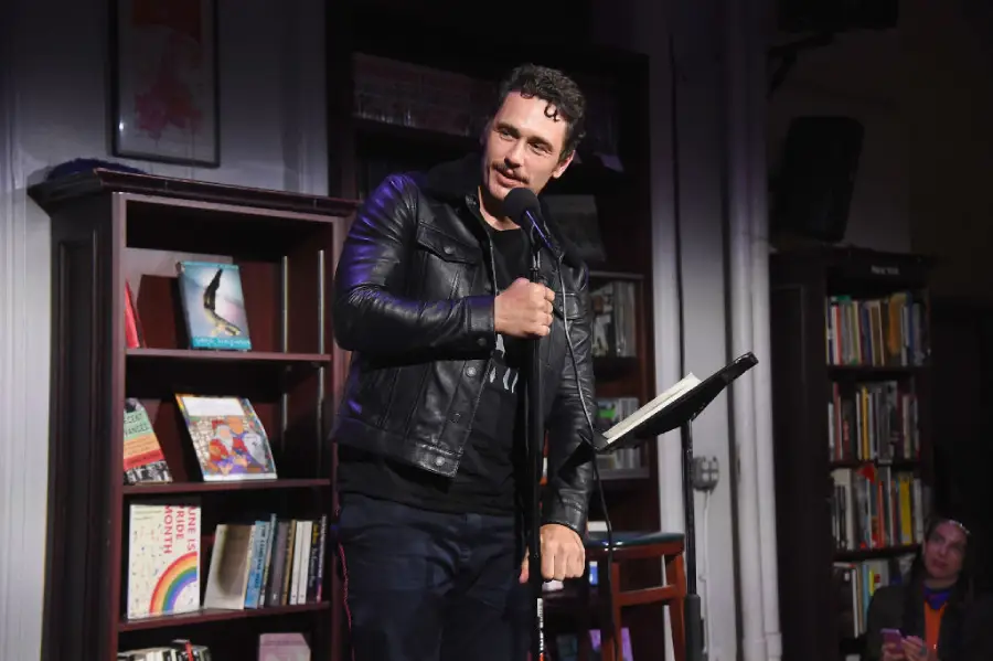 A man in a black leather jacket holds a microphone and stands on stage in a bookstore, with bookshelves and colorful books visible in the background.