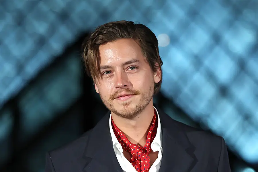 A man with light brown hair and a mustache wears a dark suit, white shirt, and red polka dot scarf, standing in front of a blurred geometric background.