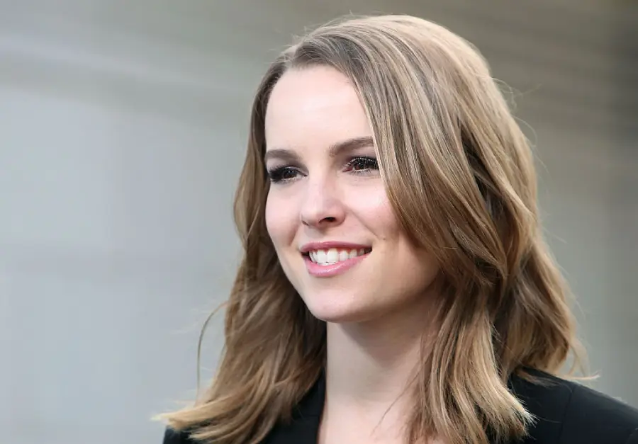 A woman with shoulder-length light brown hair smiles while looking slightly to the side. She is wearing a black top and standing in front of a blurred, neutral background.