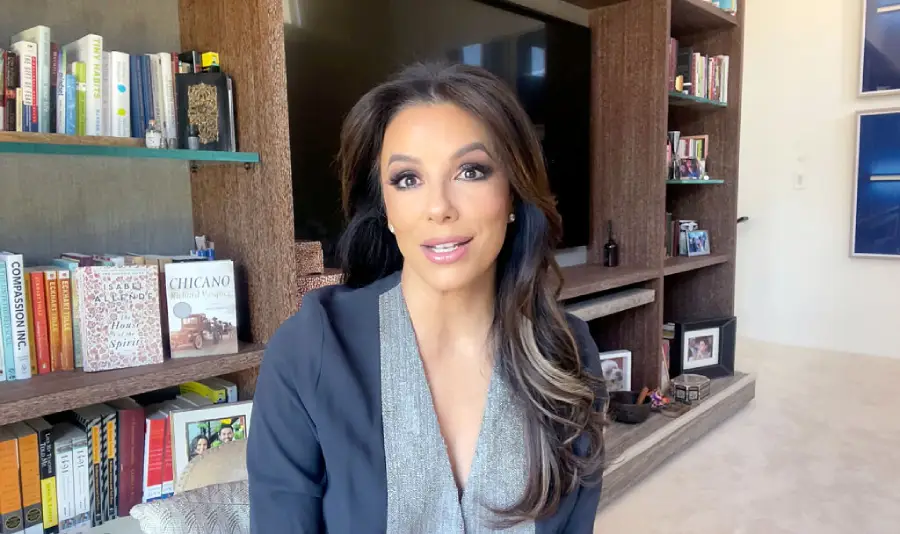A woman with long dark hair sits indoors in front of a bookshelf and television, smiling at the camera. She wears a gray top and dark blazer. The room appears well-lit and modern, with several books and photos visible.