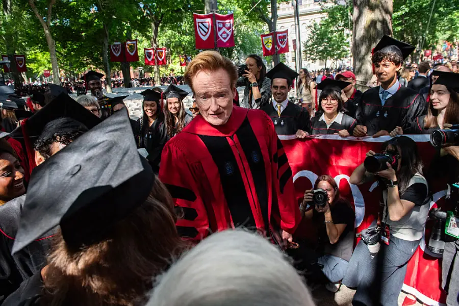 A man in a red academic robe stands among graduates in caps and gowns at an outdoor ceremony, as photographers and onlookers gather around, with trees and university banners in the background.