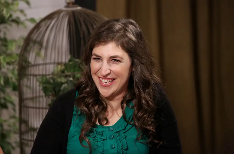 A woman with long brown hair wearing a green blouse and black cardigan smiles while sitting indoors; there is a birdcage with plants behind her.