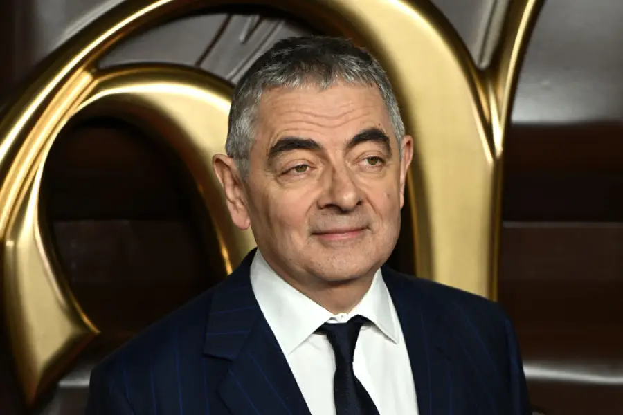 A man with short gray hair in a dark pinstripe suit and tie stands in front of a gold decorative background, smiling slightly.