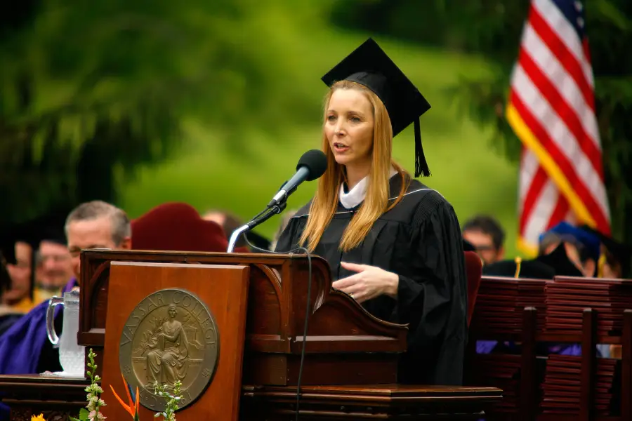 A woman in a graduation cap and gown speaks at a podium during an outdoor graduation ceremony, with people seated behind her and an American flag in the background.