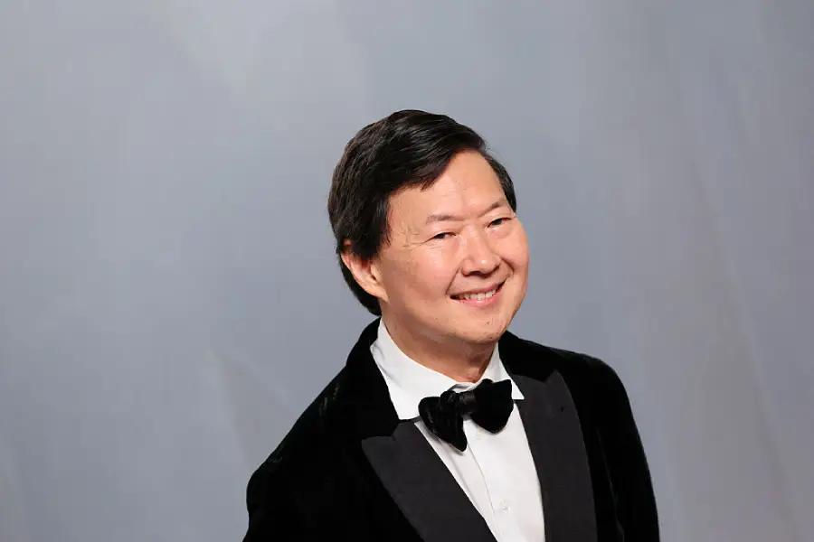 A man in a black tuxedo with a black bow tie smiles while standing against a light gray background.