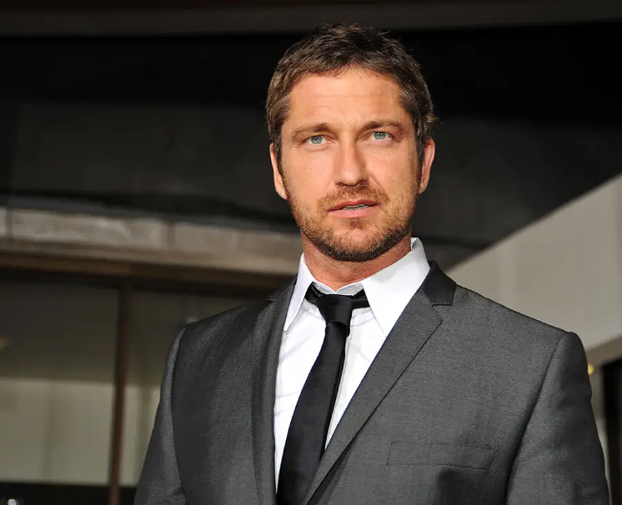 A man with short brown hair and a beard wears a dark grey suit, white shirt, and black tie, standing outdoors in front of a building, looking slightly to the side.