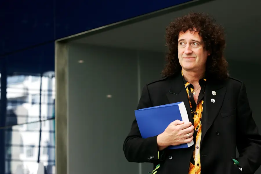 A man with curly hair in a black coat stands outside holding a blue folder, looking to the side and smiling slightly. The background features modern architecture and glass reflections.