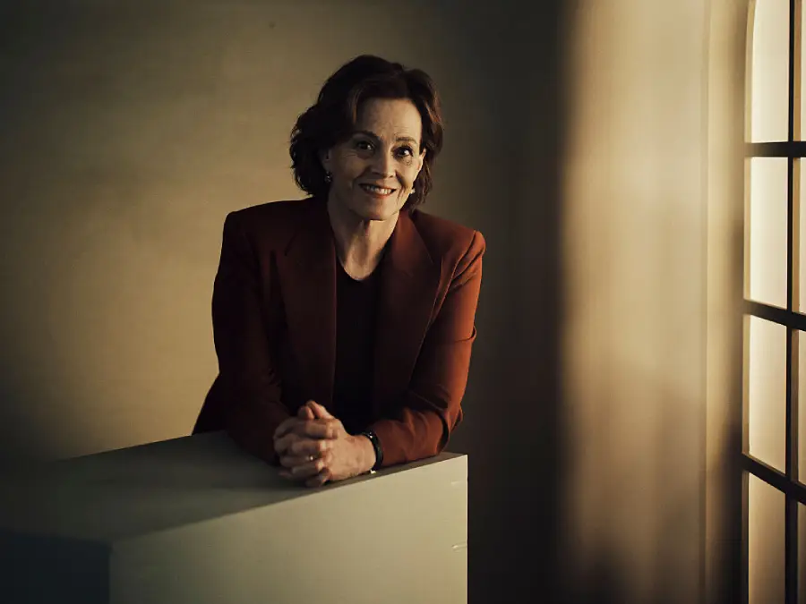 A woman with short brown hair in a red blazer leans on a white surface, smiling, in a softly lit room with light streaming through a window on the right.