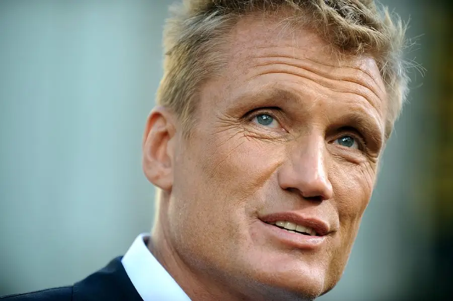 A close-up of a middle-aged man with short blond hair, blue eyes, and light skin, wearing a suit jacket and white shirt, looking slightly upward and smiling softly.