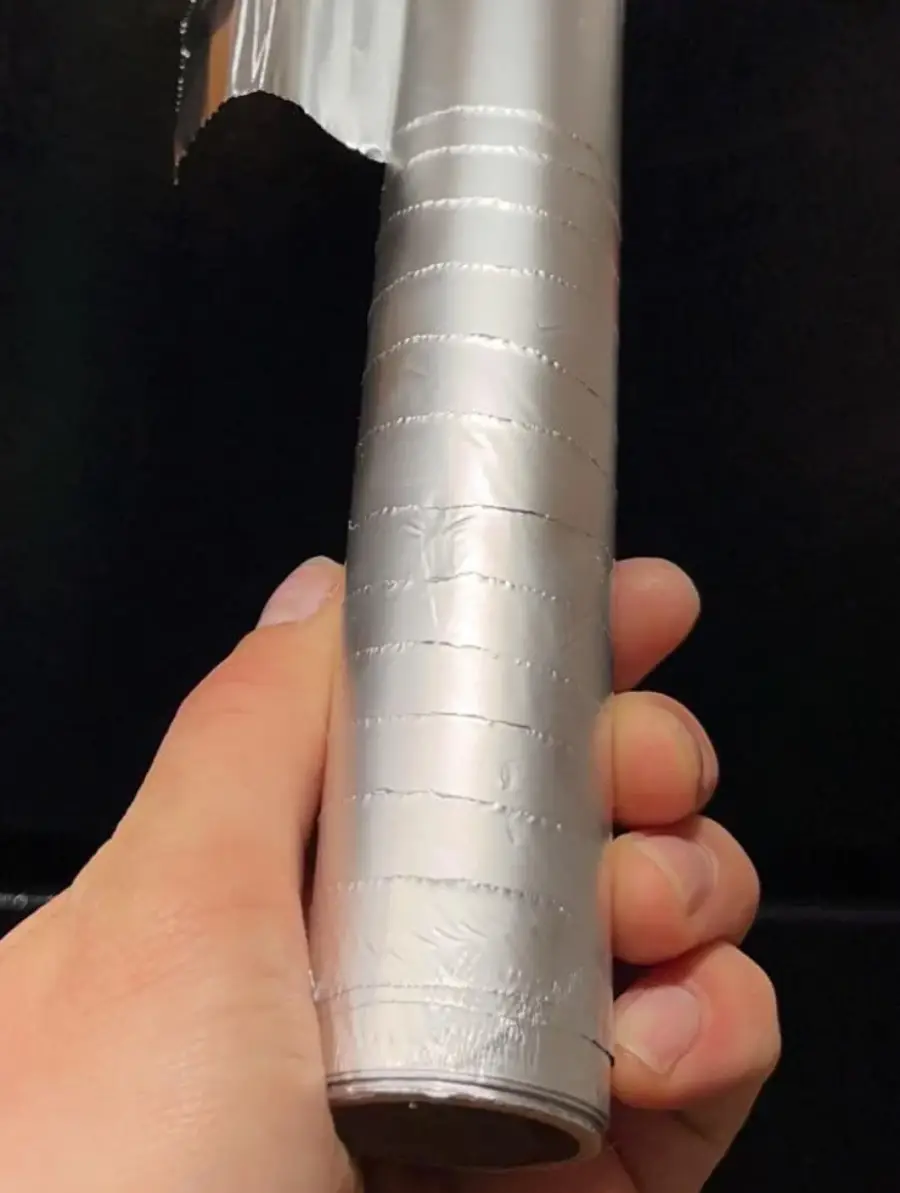 A hand holds a roll of aluminum foil with evenly spaced, straight lines running around it, making the foil appear as if it has been cleanly segmented into sections.
