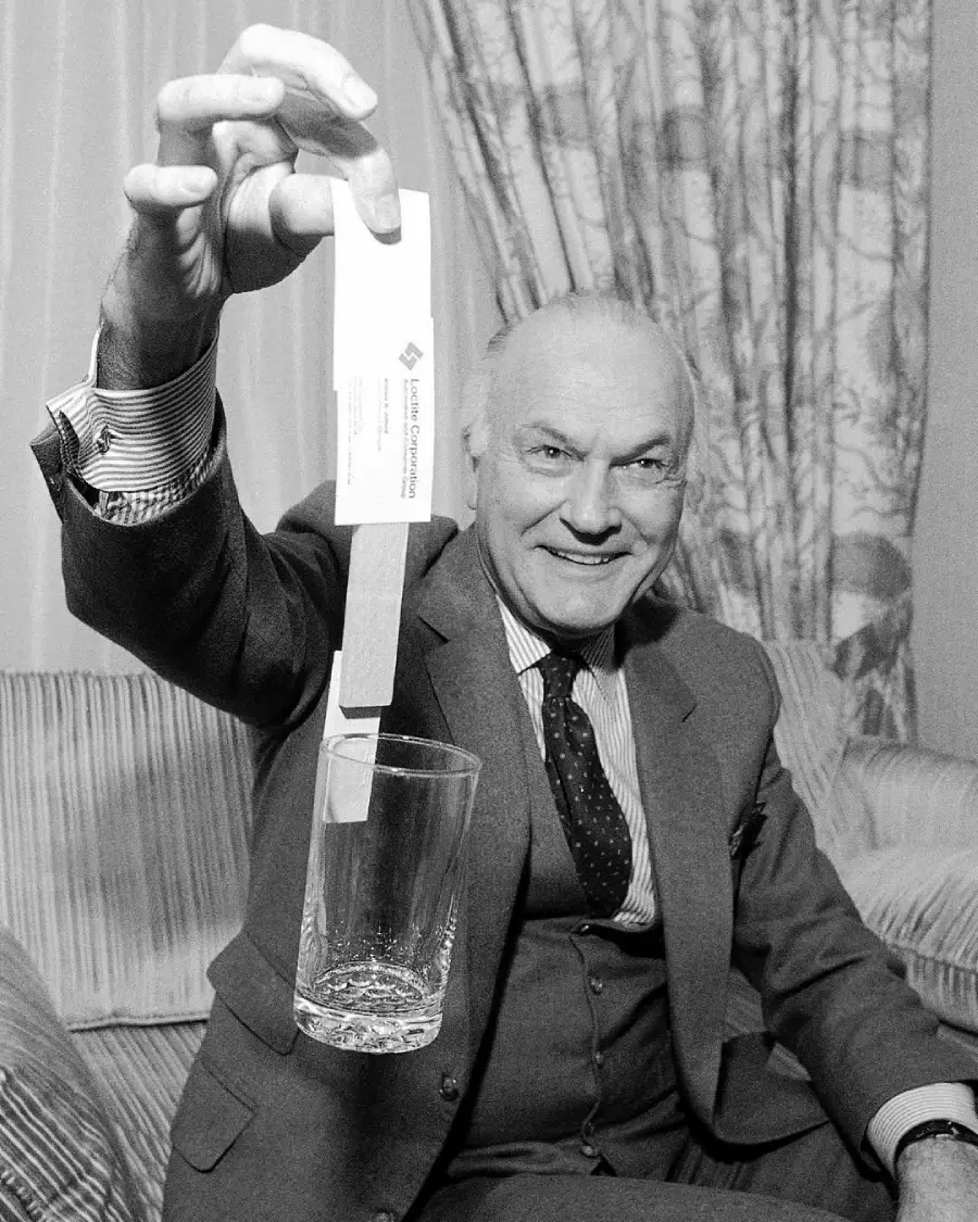A smiling man in a suit holds a business card above an empty glass while sitting on a couch with patterned curtains in the background.