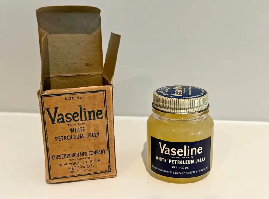 A vintage glass jar of Vaseline White Petroleum Jelly sits next to its original cardboard box on a white surface.