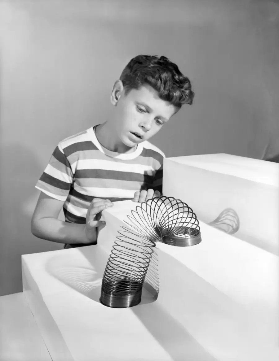 A boy in a striped shirt watches a metal Slinky toy walk down white steps, looking curious and focused.