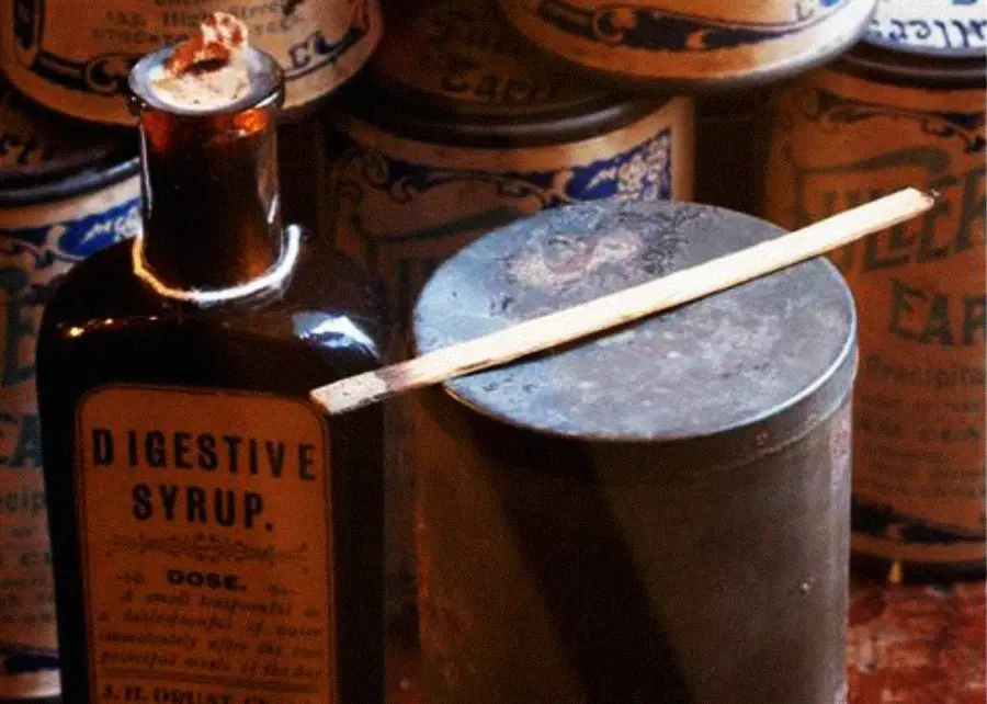 A dark glass bottle labeled “Digestive Syrup” sits next to a metal container with a wooden stick on top, surrounded by vintage medicine tins.