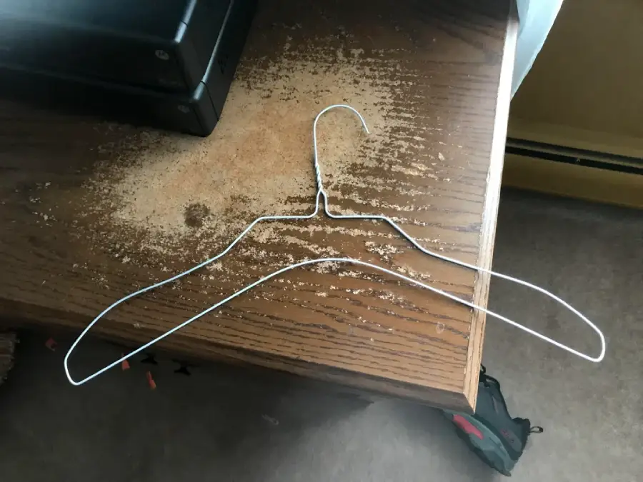 A metal wire hanger lies flat on a dusty wooden surface, with dust outlining its shape. Part of a black electronic device and a beige heater are visible in the background.