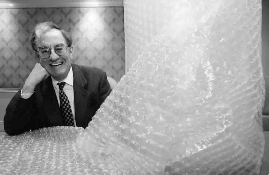 A smiling man in a suit sits beside a large sheet of bubble wrap, with patterned wallpaper in the background.