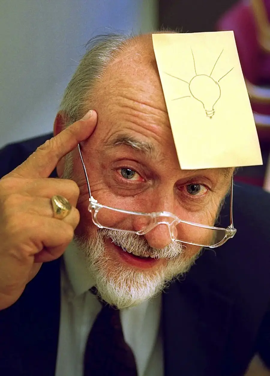 An older man with glasses and a white beard points to his head, which has a sticky note with a lightbulb drawing on his forehead, suggesting he has an idea.
