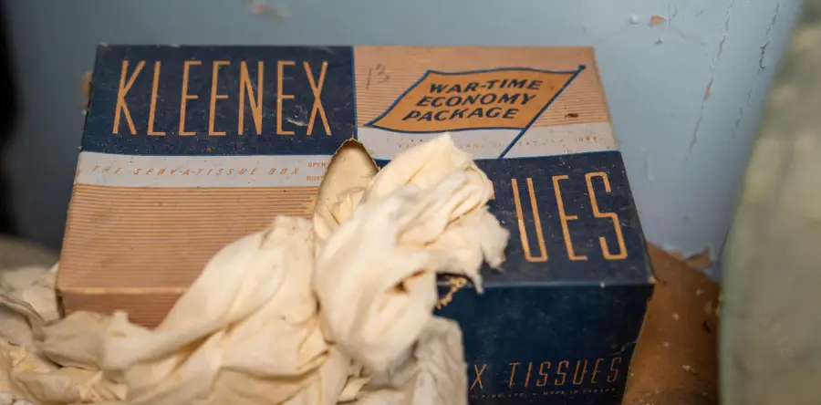 A vintage Kleenex tissue box labeled “War-Time Economy Package” sits on a dusty surface, with crumpled tissues spilling out in front.