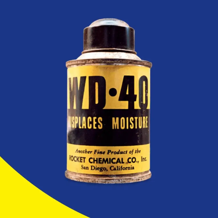 A vintage can of WD-40 with a yellow label reading “WD-40 Displaces Moisture,” made by Rocket Chemical Co., Inc., is shown against a blue and yellow background.