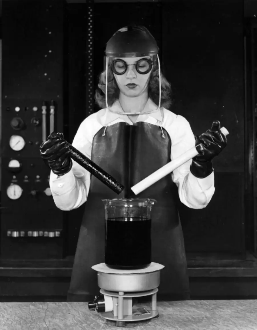 A woman in safety gear, including gloves, apron, helmet, and goggles, mixes chemicals using two rods in a beaker on a laboratory hot plate.