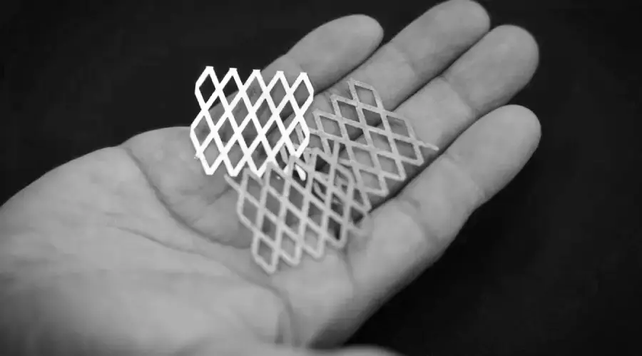 A hand holds two small, diamond-patterned mesh pieces, one light-colored and one darker, against a dark background.