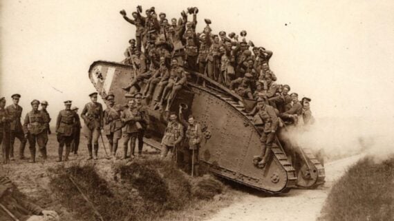 A large group of soldiers pose and cheer atop and beside a World War I-era tank on a dirt road, with some standing and others waving their hats.