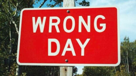 A red sign with white text reading “WRONG DAY” is mounted on a wooden post, with trees and blue sky in the background.