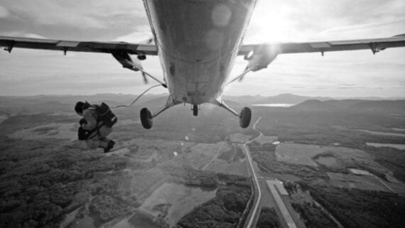 A person jumps out of a small airplane in midair, preparing to skydive over a landscape of fields, forests, and a winding road, with the sun shining in the background.