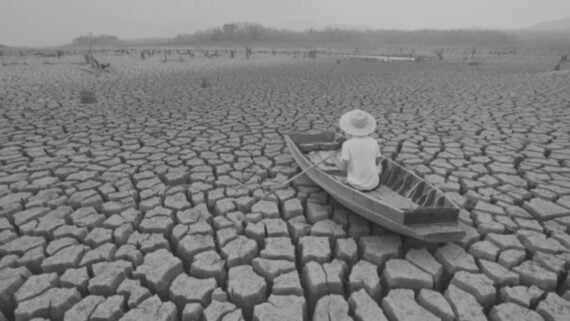 A person in a hat sits alone in a small wooden boat on a vast expanse of cracked, dry earth, highlighting drought conditions.