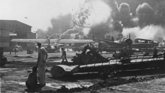 Black and white photo of a burning airfield with destroyed planes, debris, and people in naval uniforms; smoke and flames rise into the sky in the background.