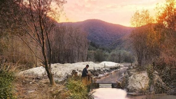 A person sits by a small stream surrounded by trees and mountains at sunset, with golden and pink hues lighting the sky and landscape.