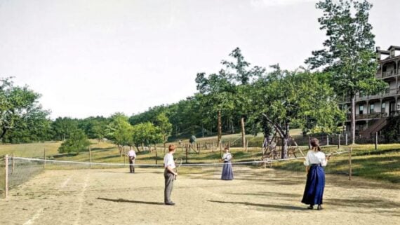 Four people play tennis on a dirt court outdoors, dressed in early 20th-century clothing. Trees and a large building are visible in the background under a clear sky.
