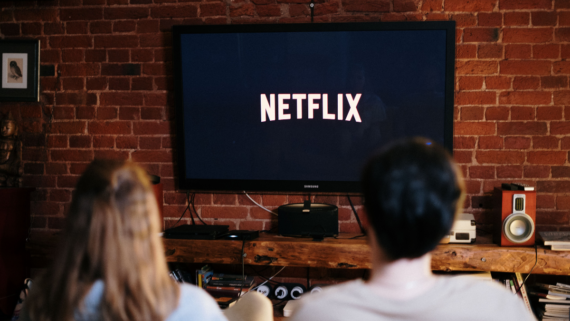 Two people sit on a couch facing a TV displaying the Netflix logo. The room has a brick wall, wooden shelf with speakers and books, and cozy decor.
