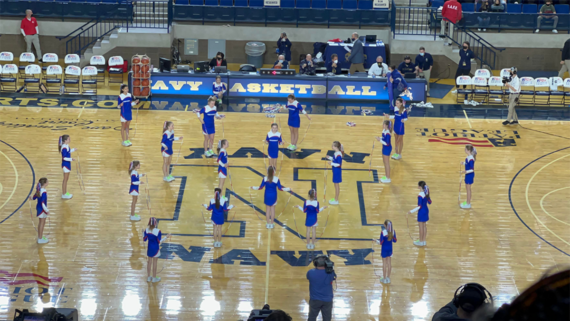 A group of cheerleaders in blue uniforms performs a routine on a basketball court with “Navy” written at center court. Spectators and staff sit in the background.
