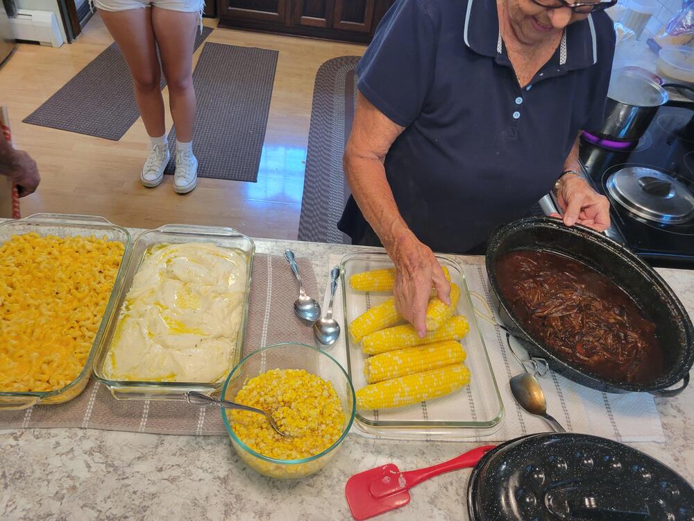 A woman serves corn on the cob from a kitchen counter with macaroni and cheese, creamy casserole, corn, and a pot of shredded meat. Another person stands in the background.